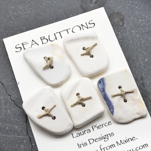 May include: Five white sea glass buttons with irregular shapes attached to a white display card labeled SEA BUTTONS, Laura Pierce, Iris Designs, and from Maine. Each button features two holes with tan string threaded through in a cross pattern.