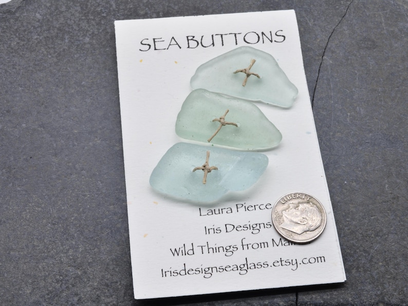 May include: Three sea glass buttons with gold-coloured metal accents are displayed on a white card. The card reads "SEA BUTTONS" at the top, with additional text below. A dime is placed next to the card for size reference.
