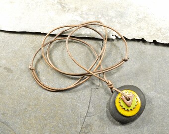 May include: A brown leather cord necklace with a round pendant. The pendant is made of a dark gray stone with a yellow and green ceramic circle on top. The ceramic circle has a copper triangle in the center.