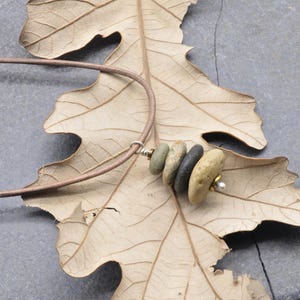 Op de afbeelding: Een handgemaakte ketting met een reeks kleine, gladde stenen in tinten groen, grijs en bruin. De stenen zijn geregen op een bruin leren koord. De ketting wordt gepresenteerd op een gedroogd blad.