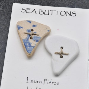 May include: Two handmade ceramic buttons. One is off-white with blue accents and the other is white. Both have a cross-stitch design and are labeled "SEA BUTTONS" and "Laura Pierce".