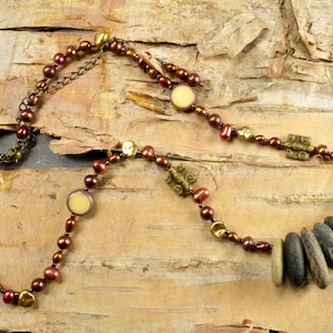 May include: A brown and gold necklace with a variety of beads, including round, oval, and rectangular shapes. The necklace also features a cluster of smooth, gray stones.