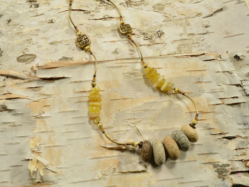 May include: A gold necklace with a variety of beads, including small round beads, larger flat beads, and a cluster of small, round, yellow beads. The necklace is on a birch bark background.
