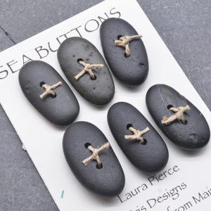 May include: Six oval-shaped dark gray stone buttons arranged on a white display card labeled SEA BUTTONS. Each button features two drilled holes with light tan twine threaded through in an X pattern. The card includes the text Laura Pierce.