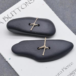 May include: Two dark gray, pebble-shaped buttons with natural twine accents. The buttons are on a white card with the text "SEA BUTTONS" printed in black. The buttons have two holes for sewing.