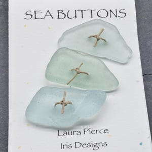 May include: Three sea glass buttons in shades of blue and green, each with a tan cross-shaped embellishment. The card reads "SEA BUTTONS" at the top, with "Laura Pierce Iris Designs" at the bottom. The buttons are on a white card.
