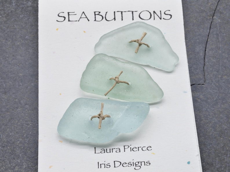 May include: Three sea glass buttons in shades of blue and green, each with a tan cross-shaped embellishment. The card reads "SEA BUTTONS" at the top, with "Laura Pierce Iris Designs" at the bottom. The buttons are on a white card.