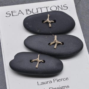 May include: Three dark gray, oval-shaped buttons with a cross-shaped, tan thread detail. The buttons are on a white card that reads "SEA BUTTONS" at the top. The card also includes the name "Laura Pierce Designs".