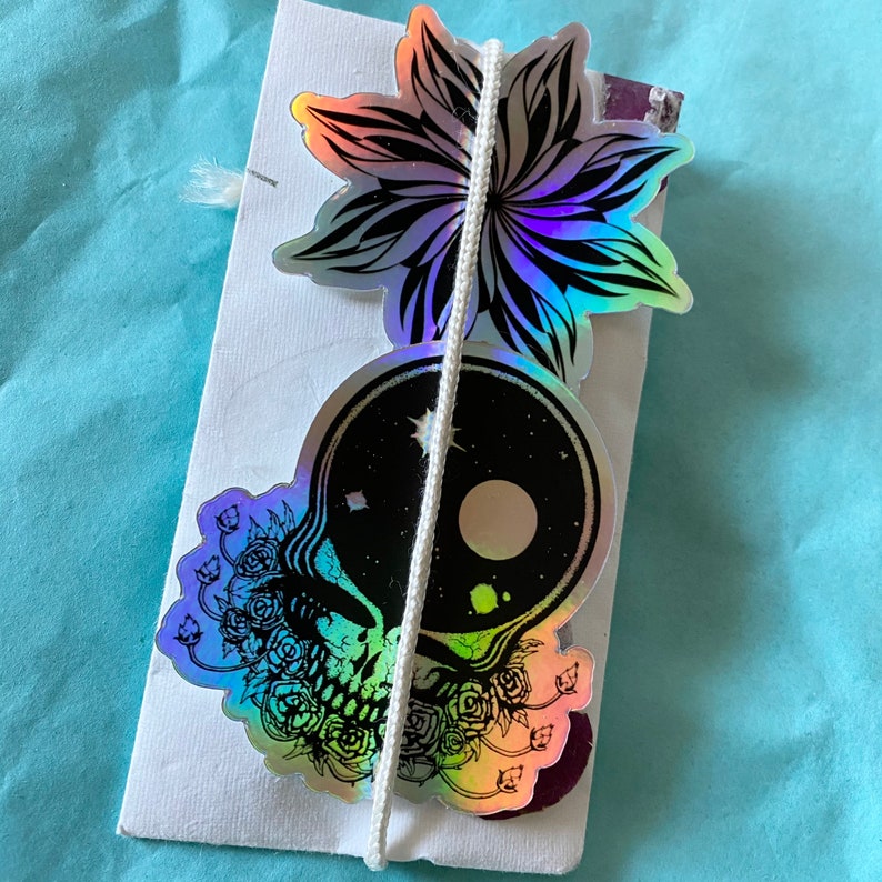 May include: A white package tied with white string, featuring holographic stickers. The stickers include a flower, a skull with roses, and a circular design with a galaxy theme. The background is a light blue surface.