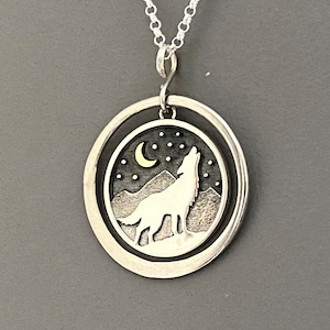 May include: Silver-tone pendant necklace featuring a howling wolf silhouette against a night sky with a crescent moon and stars. The pendant is circular with a double-layered design, and hangs from a delicate chain.