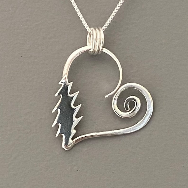 May include: A silver pendant necklace featuring a heart-shaped design with a stylized tree on one side and a spiral on the other. The pendant hangs from a delicate chain, showcasing a nature-inspired theme.
