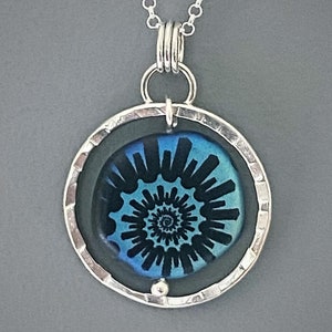 May include: A silver pendant necklace featuring a circular pendant with a blue and black spiral design. The pendant is encased in a hammered silver frame and hangs from a delicate silver chain. The design is reminiscent of a nautilus shell.