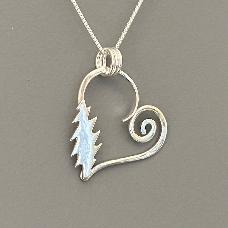 May include: A silver pendant necklace featuring a heart-shaped design. The pendant has a stylized tree on one side and a spiral on the other. The necklace chain is a delicate silver color.