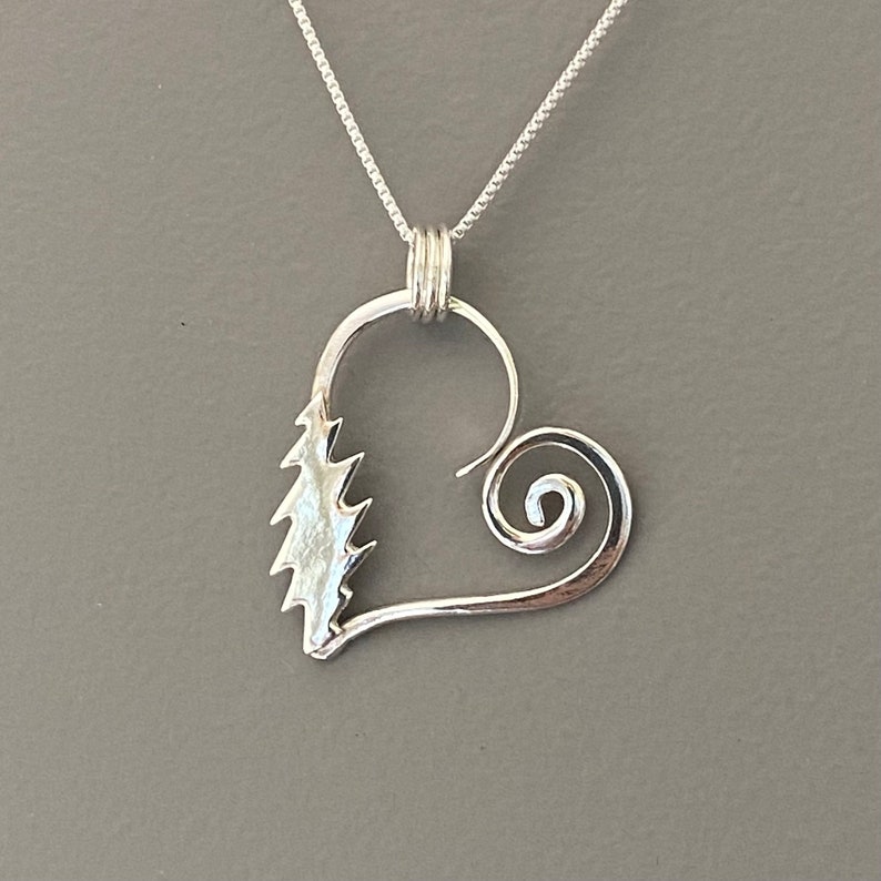 May include: A silver pendant necklace featuring a heart-shaped design. The pendant has a stylized tree on one side and a spiral on the other. The necklace chain is a delicate silver chain.