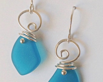 May include: A pair of handcrafted earrings featuring teardrop-shaped, light blue sea glass pendants. The pendants are accented with silver wire wrapping and a decorative swirl at the top, suspended from silver ear wires. A simple and elegant jewelry design.