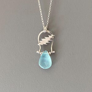 May include: A silver necklace featuring a teardrop-shaped, light blue gemstone pendant. The pendant is suspended from a silver bail with a lightning bolt design. The necklace has a delicate silver chain.