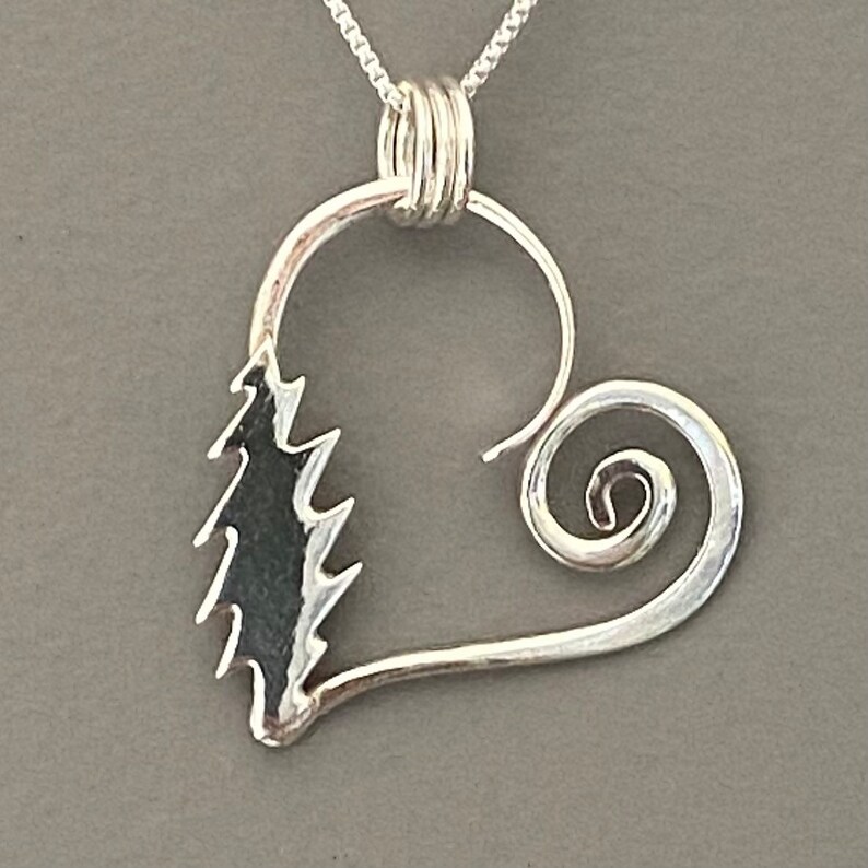May include: A silver pendant necklace featuring a heart-shaped design. The pendant has a stylized tree on one side and a spiral on the other. The necklace chain is a delicate silver color.