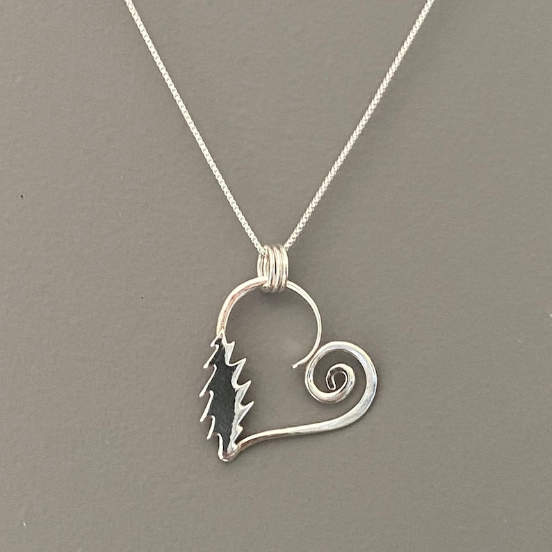 May include: A silver pendant necklace featuring a heart-shaped design. The pendant has a stylized tree on one side and a spiral on the other. The necklace chain is a delicate silver chain.