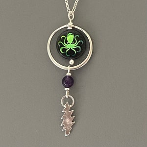 May include: Silver necklace with a circular pendant featuring a green octopus design on a black background. Below the pendant is a purple bead and a silver lightning bolt charm. The necklace hangs on a silver chain.