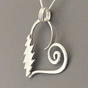May include: A silver pendant necklace featuring a heart-shaped design. The pendant has a stylized tree on one side and a spiral on the other. The necklace hangs on a delicate silver chain, perfect for everyday wear.
