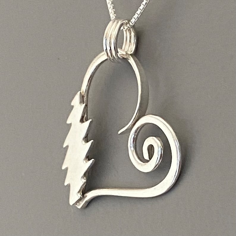 May include: A silver pendant necklace featuring a heart-shaped design. The pendant has a stylized tree on one side and a spiral on the other. The necklace hangs on a delicate silver chain, perfect for everyday wear.