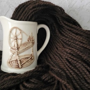 May include: A white ceramic pitcher with a brown spinning wheel design. The pitcher is surrounded by a pile of dark brown yarn.