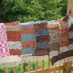 May include: Five woven rugs with fringe edges in various colors and patterns, including pink, red, blue, and green, hanging on a wooden railing.