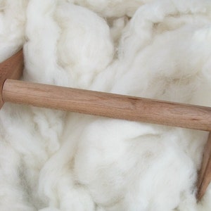 May include: A wooden spinning wheel with a natural finish. The wheel is made of two curved pieces of wood that are connected by a wooden dowel. The wheel is sitting on a bed of white wool.