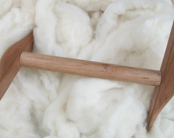 May include: A wooden spinning wheel with a natural finish. The wheel is made of two curved pieces of wood that are connected by a wooden dowel. The wheel is sitting on a bed of white wool.