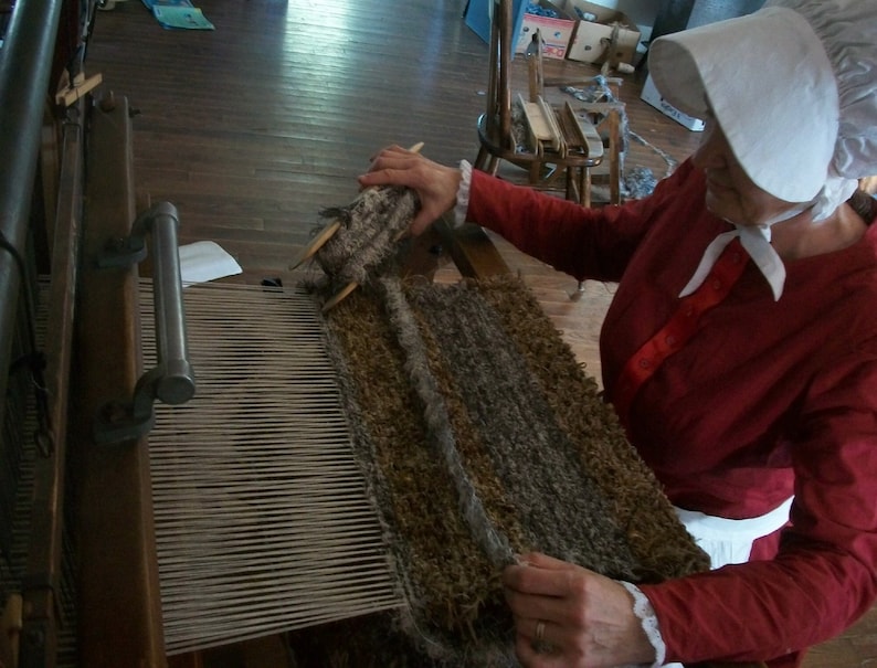 May include: A woman in a red dress and white bonnet is weaving a rug on a loom. The rug is made of brown and gray wool and is being woven with a white thread.