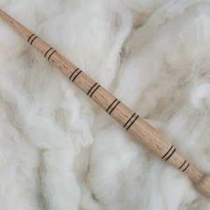 May include: A wooden spindle with a pointed tip and a dark brown stripe design. The spindle is lying on a white fluffy surface.