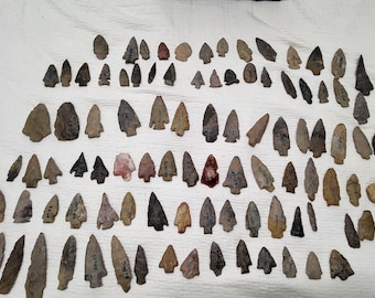 May include: A large collection of stone arrowheads and projectile points in various shades of gray, brown, tan, and black. The items are arranged in rows, featuring diverse triangular and leaf-shaped silhouettes with pointed tips and notched bases.