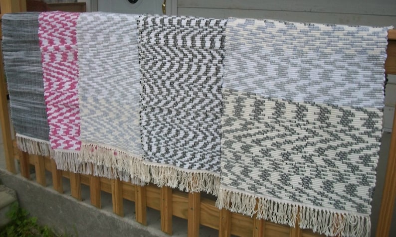 May include: Four woven rugs with fringe edges. The rugs are in various patterns and colors, including gray, white, pink, and black.