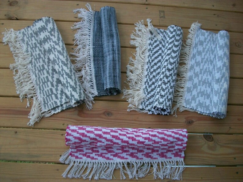 May include: Five woven rugs with fringe edges in various patterns. The rugs are in shades of gray, white, and pink. The rugs are laid out on a wooden surface.