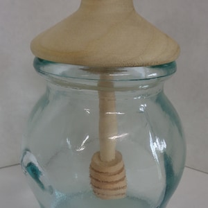 May include: A clear glass honey jar with a wooden lid and a wooden honey dipper inside. The jar has a textured surface and a rounded shape.