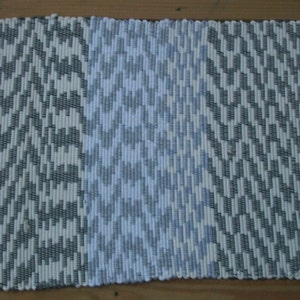 May include: A white and gray woven rug with a geometric pattern. The rug has a fringe border on all sides.