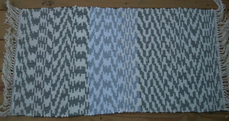 May include: A white and gray woven rug with a geometric pattern. The rug has a fringe border on all sides.