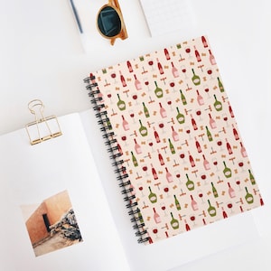 Puede incluir: Una libreta de espiral con una cubierta de color crema que presenta un patrón repetitivo de botellas de vino, vasos y sacacorchos en tonos rojos, rosas, verdes y marrones.