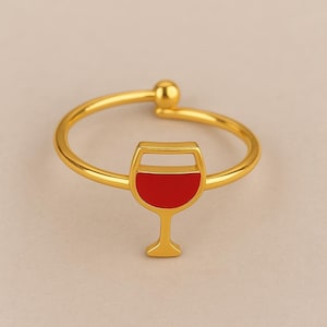 Può includere: Un anello dorato con un design a calice di vino rosso. L'anello ha una fascia semplice con un piccolo dettaglio a sfera nella parte superiore. Il calice è riempito di smalto rosso.