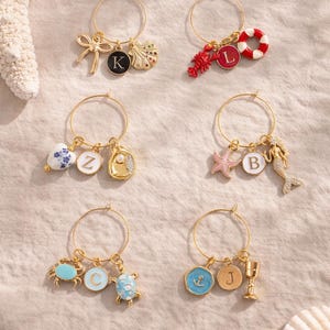 May include: Gold hoop earrings with assorted charms: a bow, shell, lobster, life ring, starfish, mermaid, crab, turtle, anchor, and chalice. Each earring has a letter charm.
