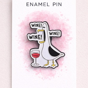 May include: Enamel pin featuring a cartoon seagull with a wine glass and speech bubbles that say "WINE!". The pin is white, black, and yellow, with a pink watercolor background and the text "ENAMEL PIN" at the top.