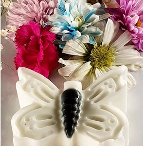 Peut inclure: Un savon blanc en forme de papillon avec un centre noir, sur fond de fleurs colorées. Les fleurs comprennent des fleurs roses, bleues, blanches et violettes, créant un contraste vibrant.