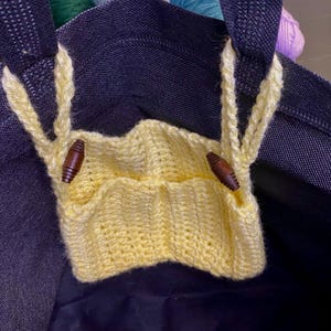 May include: A navy blue tote bag with a yellow knitted pocket. The pocket is secured with two wooden toggle buttons. The bag has yellow knitted handles and is filled with colourful yarn.