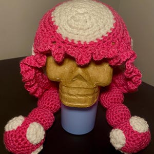 May include: A handmade crochet hat in bright pink and white, resembling a mushroom. The hat features a white circle on top, a scalloped brim, and two long, beaded strands with white polka dots. The hat is displayed on a mannequin head.