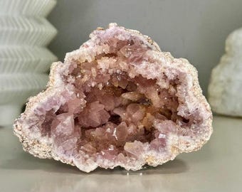May include: A raw amethyst geode featuring a cluster of pale pink and light purple crystalline points within a rough, beige-colored rocky exterior. The interior cavity is filled with jagged, translucent crystal formations that catch the light.