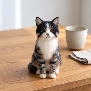 May include: A needle-felted cat figurine featuring a black, brown and white tabby coat pattern. The cat has a white chest, dark ears and realistic dark eyes, sitting in an upright position with its front paws placed together on a wooden surface.
