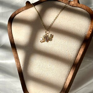 Può includere: Una collana color oro con un ciondolo a farfalla con dettagli rosa, verdi e trasparenti e una piccola perla. La collana è esposta su un espositore di gioielli in legno con un retro in tessuto color crema.