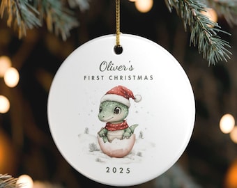 Könnte beinhalten: Runder, weißer Weihnachtsschmuck mit einer Illustration eines Baby-Dinosauriers mit Weihnachtsmütze und Schal, der aus einem Ei schlüpft. Der Text "Oliver's First Christmas" und das Jahr "2025" sind auf dem Ornament aufgedruckt.