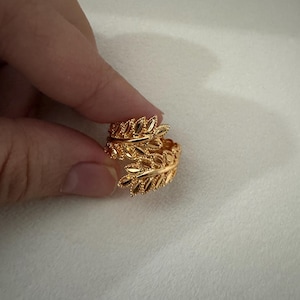 May include: A gold-colored ring with a leaf design. The ring wraps around the finger with detailed leaf patterns. The ring is held by a hand against a white background. The ring is likely made of metal.