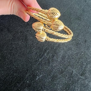 May include: Gold-colored bracelets with a twisted rope design and textured ball accents. The bracelets are held in a hand against a dark background. The jewelry appears to be made of metal.